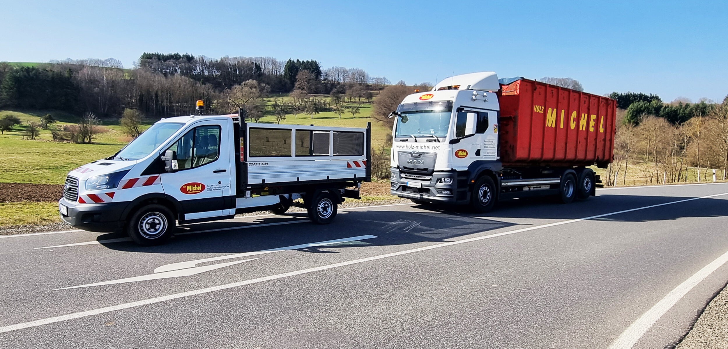 Ein weißer Kleintransporter und ein Liefer-Lkw mit einem auffälligen roten Container stehen hintereinander am Rand einer asphaltierten Landstraße.